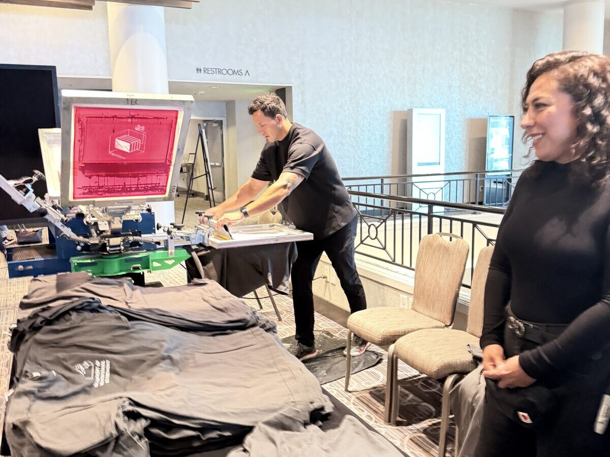 Guests in suits gathered around a press table at a corporate event, with stacks of folded navy and maroon printed event tees in the foreground