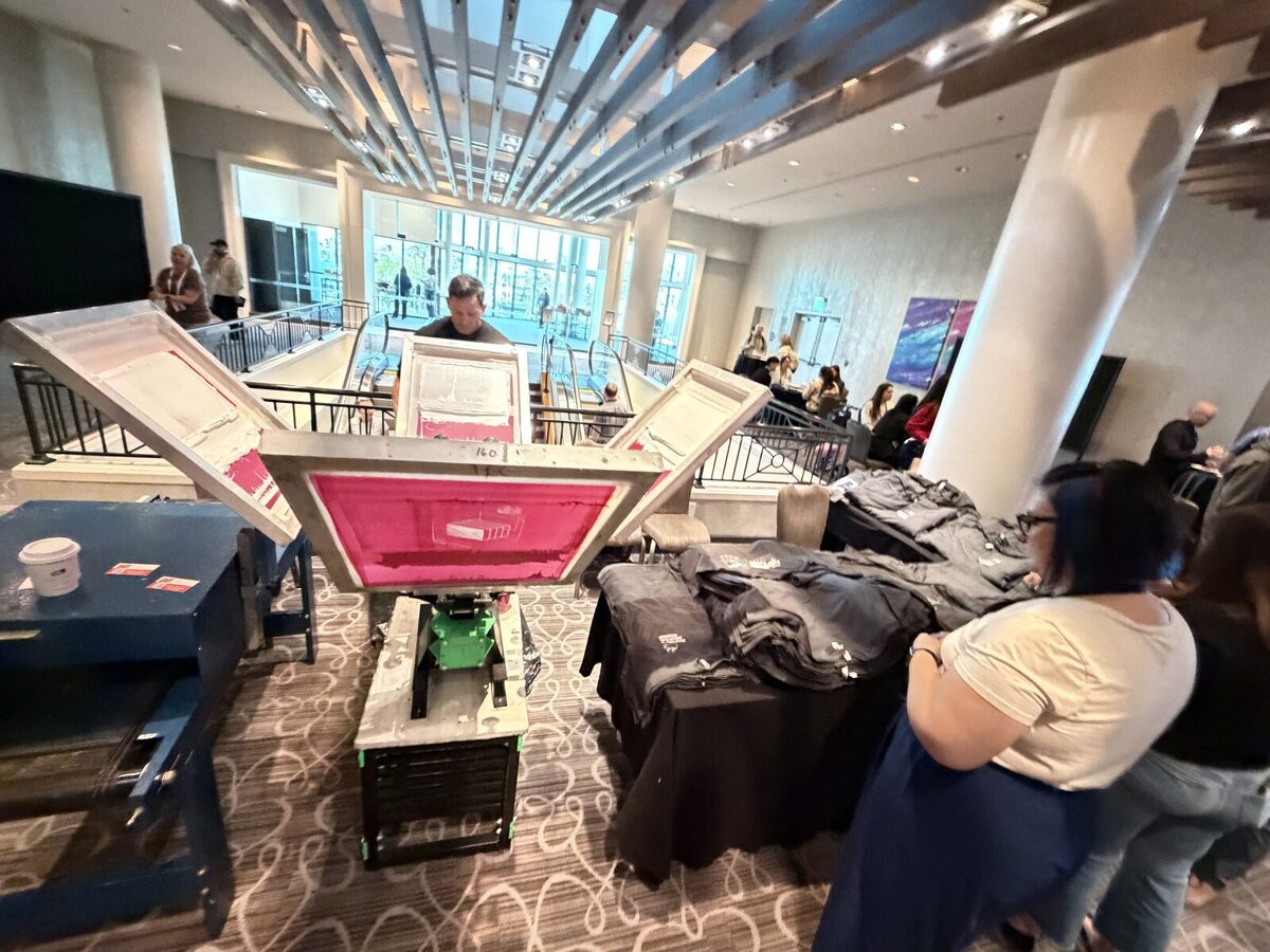 A printer pulling a pink-lit screen on a press at a conference, with folded printed tees on the table and a guest watching beside large windows