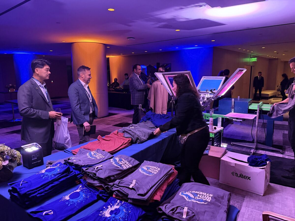 Guests in suits gathered around a press table at a corporate event, with stacks of folded navy and maroon printed event tees in the foreground