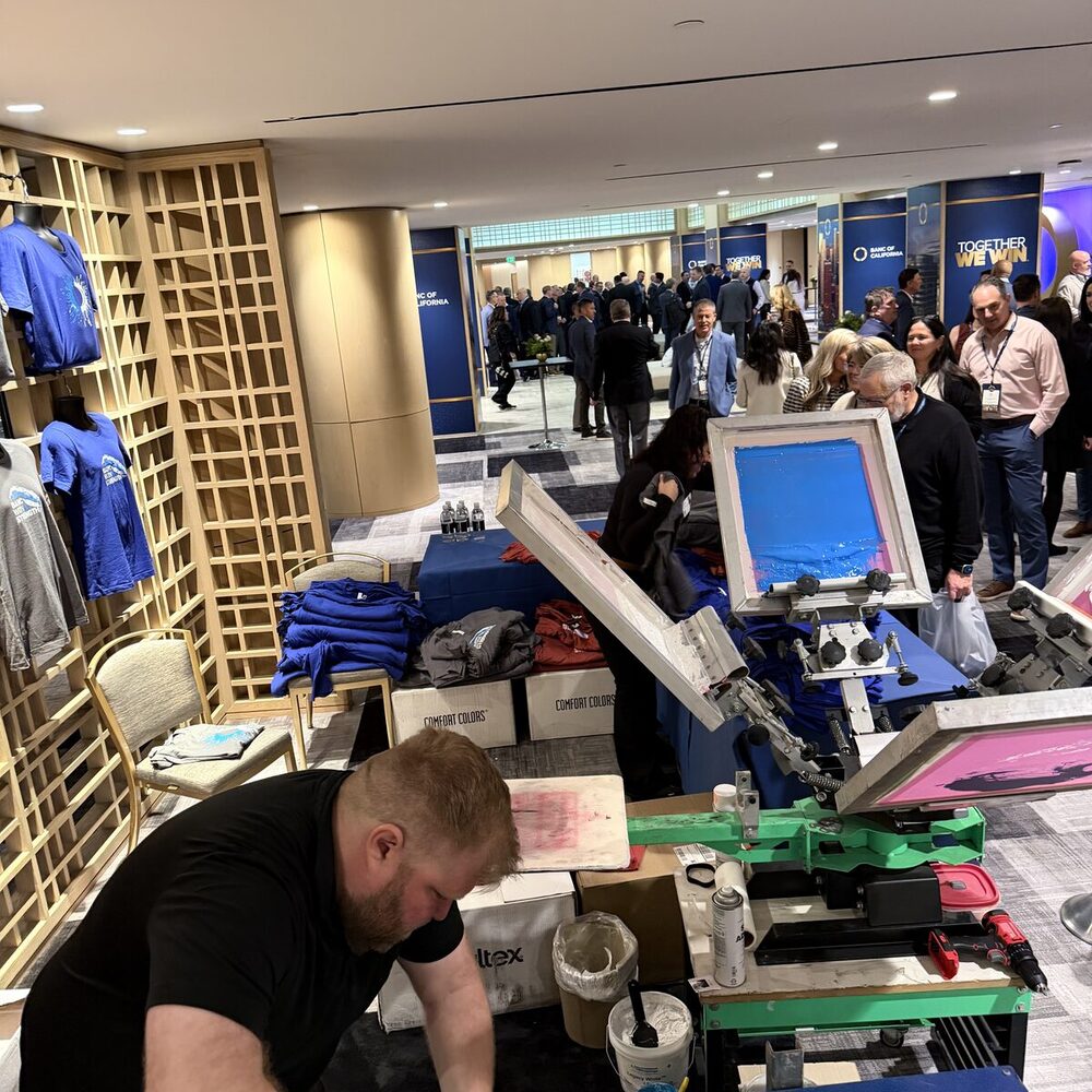 A printer loading a screen on a press at a corporate reception, with finished blue event tees displayed on a rack and a crowd of attendees behind