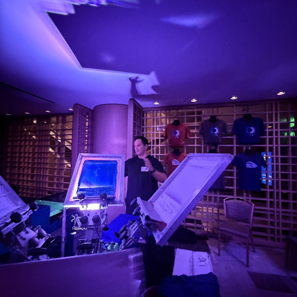 A printer beside a press under blue light, with finished orange, gray, and blue starburst-print tees displayed on the wall behind
