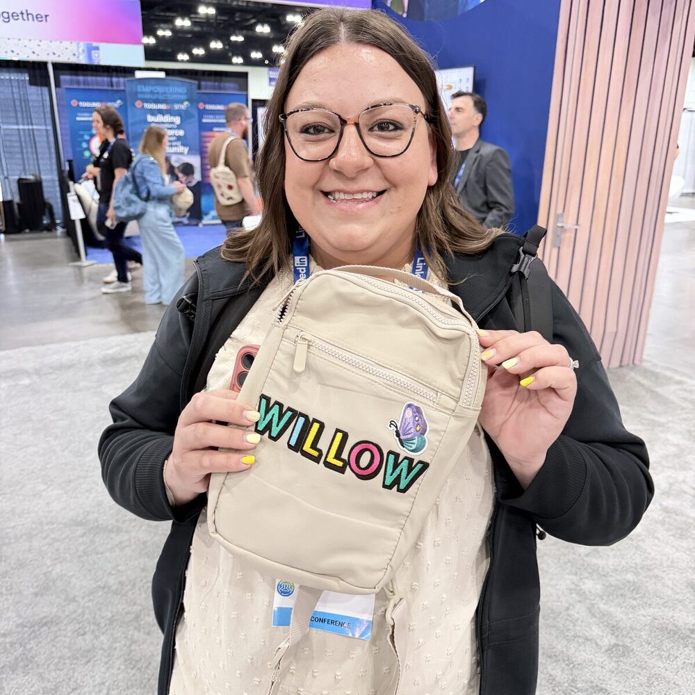A smiling guest holding up a tan crossbody bag personalized on-site with the embroidered name WILLOW in colorful letters and a small butterfly