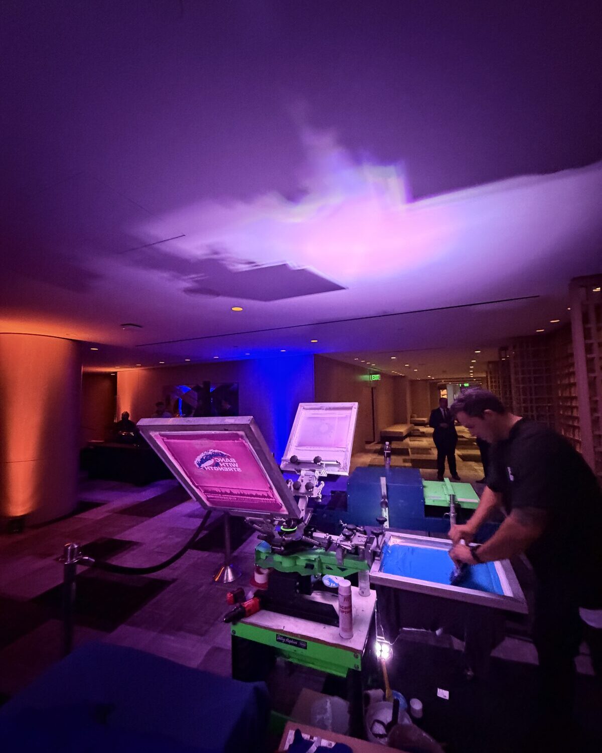 A Merch Troop printer pulling ink across a screen on a manual press at an evening event, lit in deep purple and blue, a magenta-lit logo screen on the platen