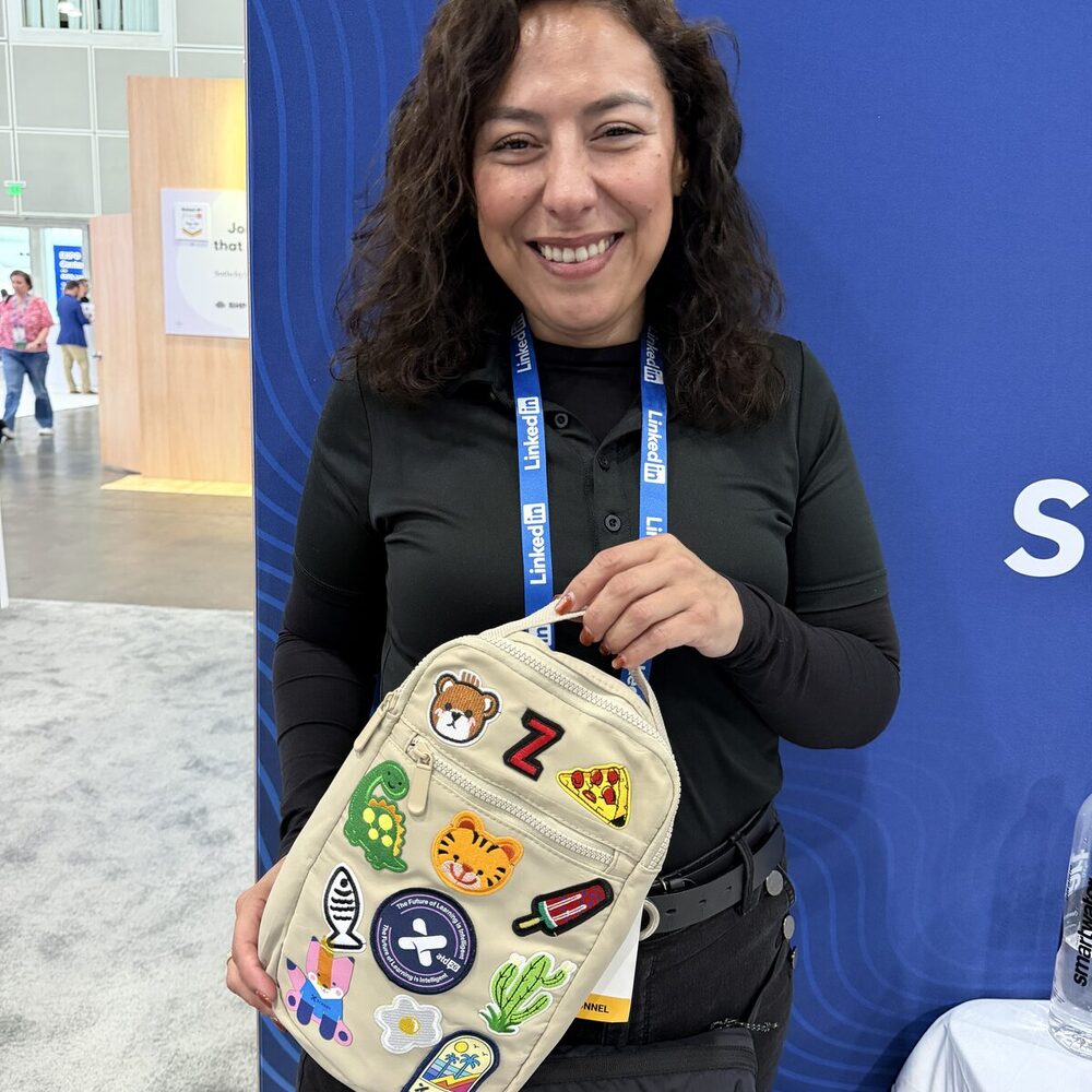 A smiling guest holding a tan bag covered in custom embroidered patches — a bear, pizza slice, dinosaur, cactus, and more