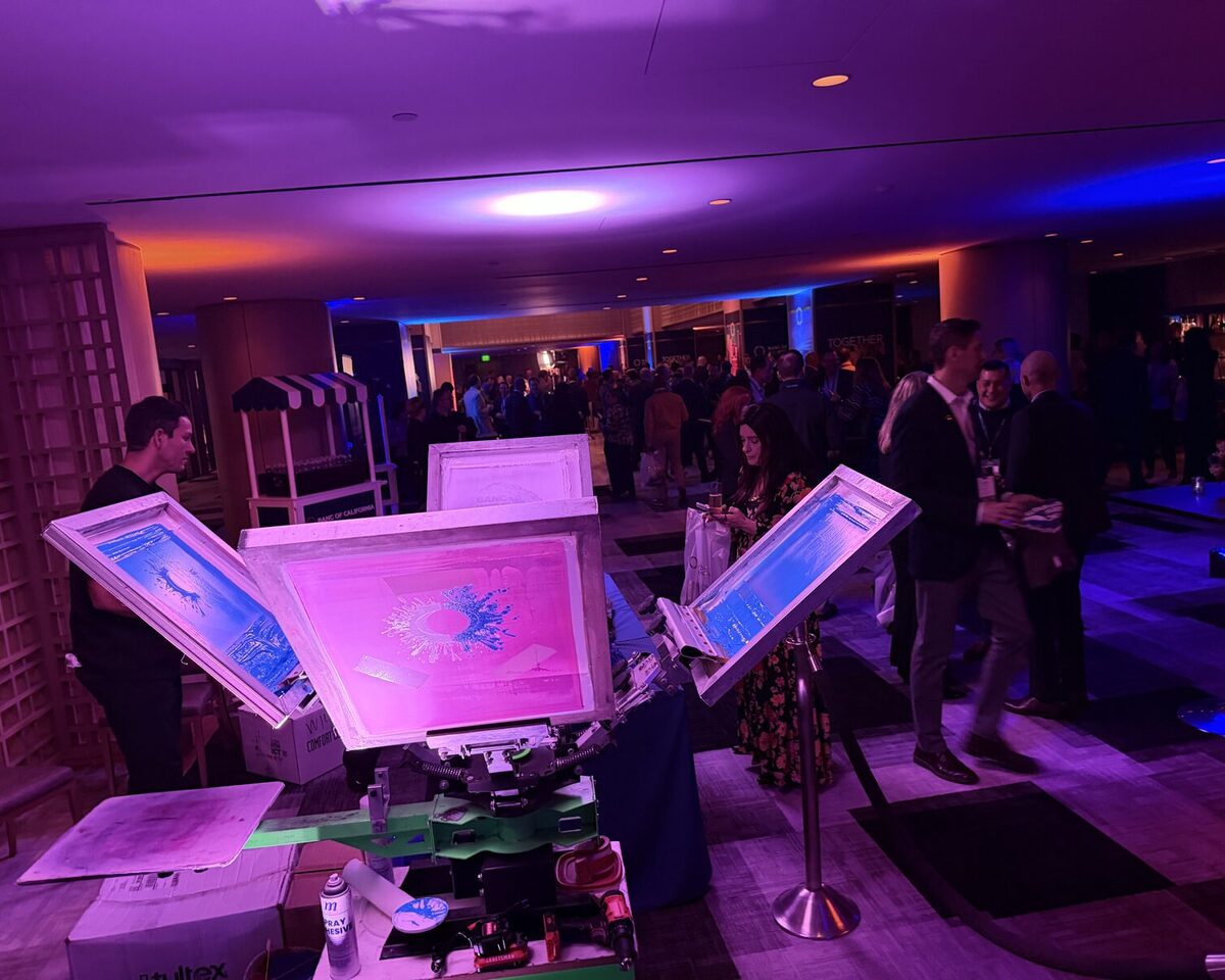 A carousel screen-printing press with its screens splayed open and a pink-lit screen on the platen, event guests mingling in the background
